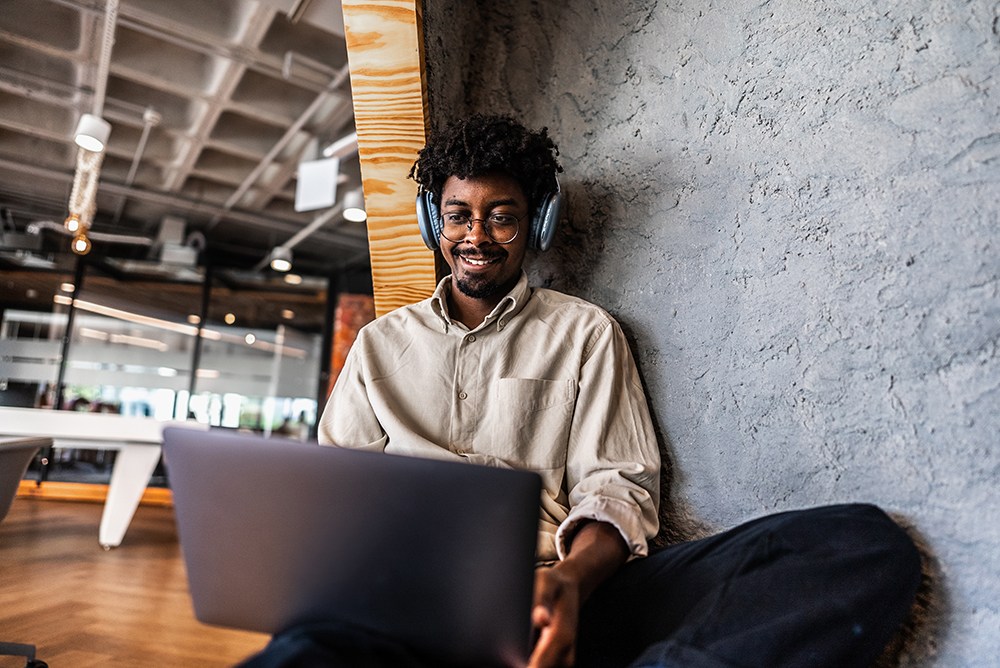 A person is sitting on the floor against a stone wall, wearing headphones and smiling while using a laptop. The background features an open office space with large windows and modern furniture.