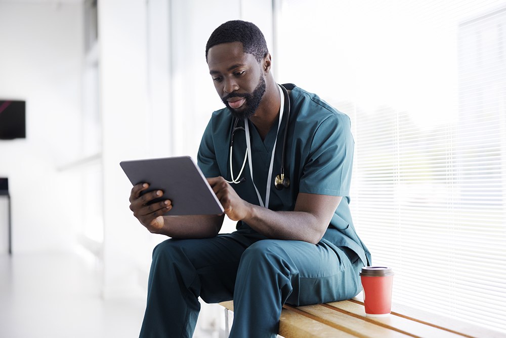 A healthcare professional in teal scrubs sits on a wooden bench with a stethoscope around his neck. He is focused on a tablet, with a red disposable cup nearby. Sunlight streams through the window blinds behind him.