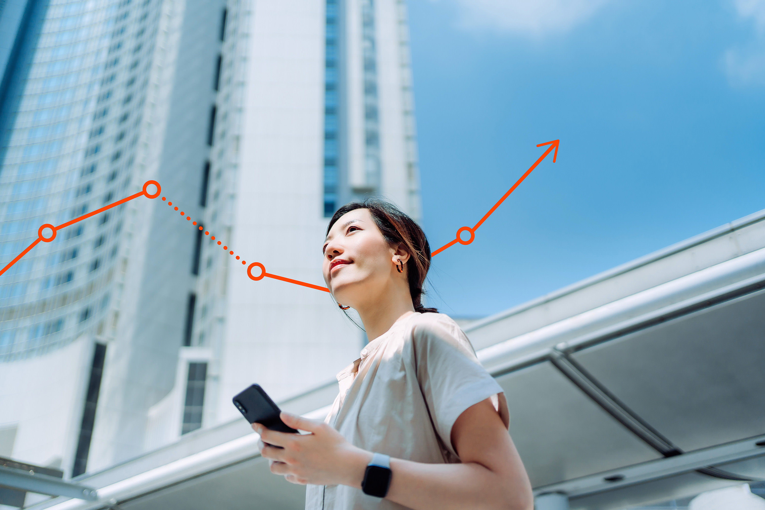 view upward at smiling woman, holding her phone, standing in front of a modern skyscraper in a city against a blue sky