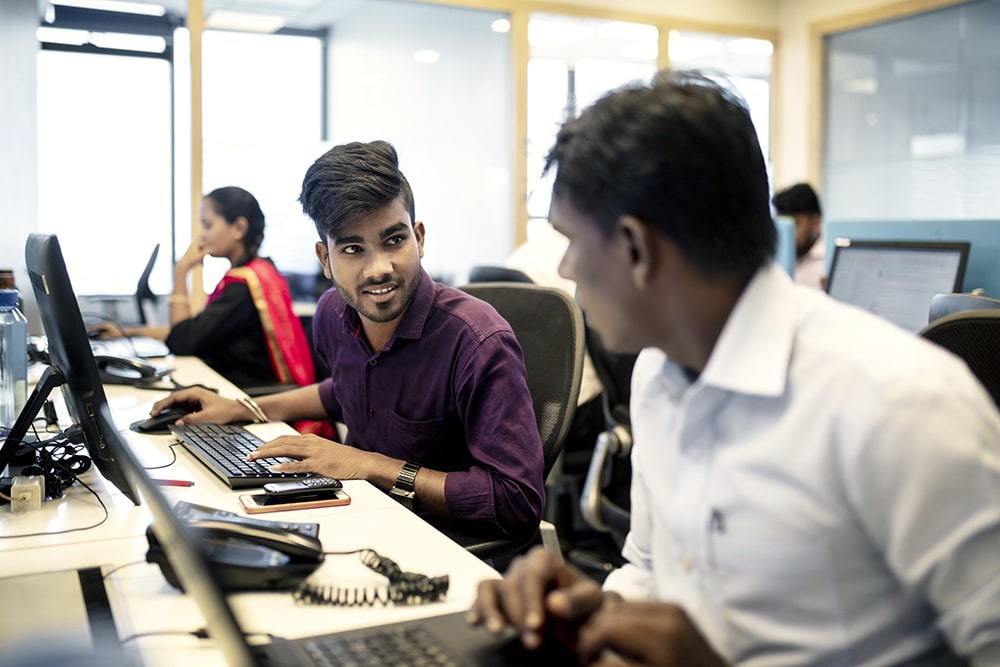 two Indian men working together at a long desk with a woman in the background