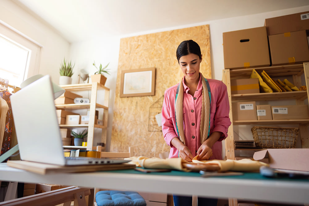 A woman wearing a pink shirt and scarf stands at a table wrapping items in a small workspace filled with shelves, boxes, plants, and a laptop. The room is well-lit with natural light coming from a window on the left.