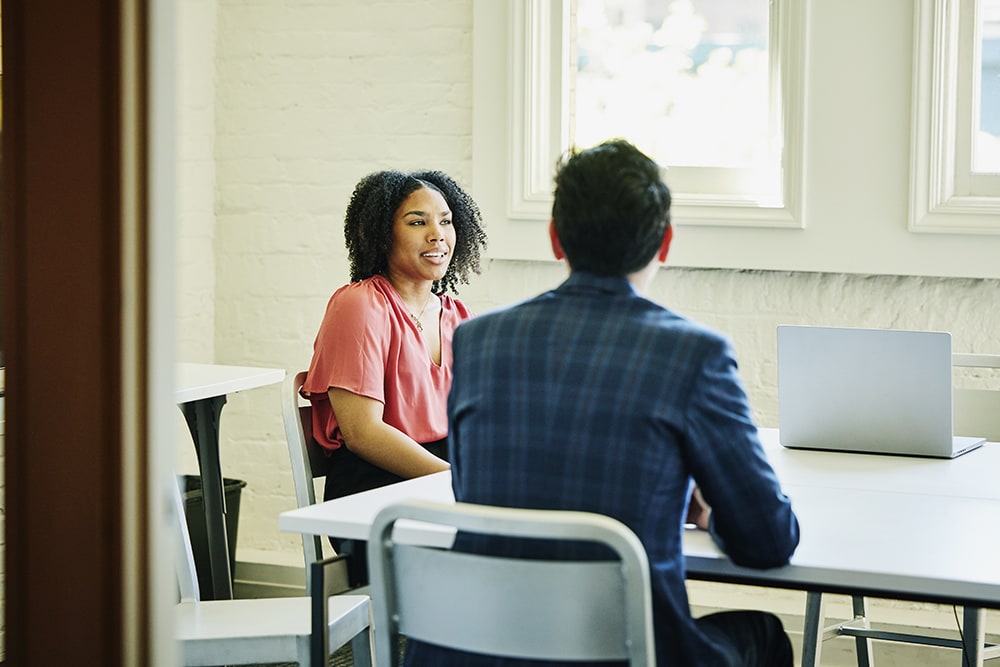 A woman and a man are sitting at a table in an office setting, having a discussion. A laptop is open on the table in front of them. The room has white walls and large windows letting in natural light.