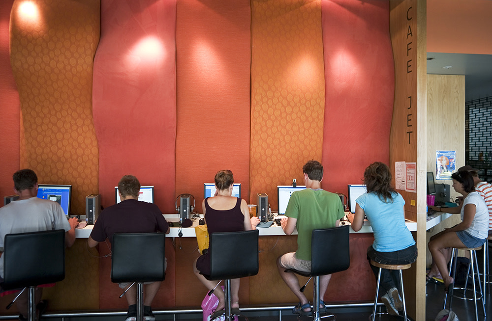 People sit on stools using computers at a counter. The background features a red and orange wall with geometric patterns. A sign on the side reads Cafe Jet.