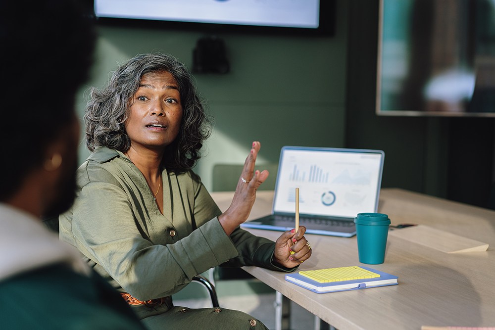 A person with gray hair gestures while speaking during a meeting. Theyre seated at a table with a notebook, blue coffee cup, and an open laptop displaying charts. Another person is partially visible in the foreground, listening attentively.