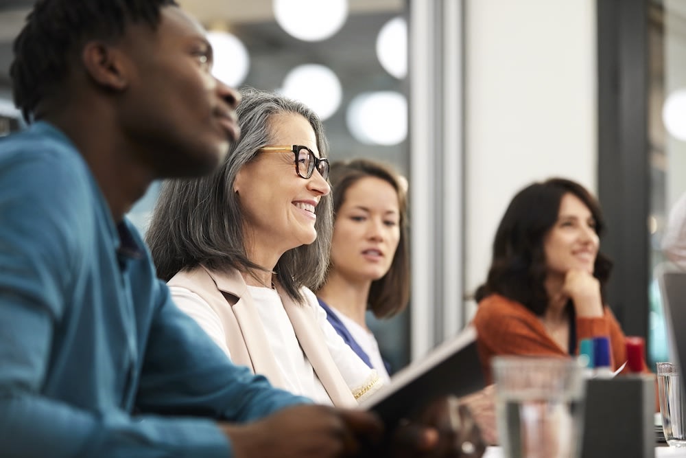 Four people, diverse in age and gender, sit side by side at a table in a bright office, attentively watching and smiling at something out of view. Papers, pens, and glasses of water are on the table in front of them.