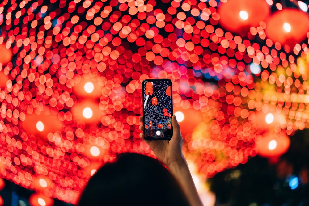 A person holds up a smartphone to capture a photo of vibrant red lanterns and lights, which create a bokeh effect in the background during a night festival.