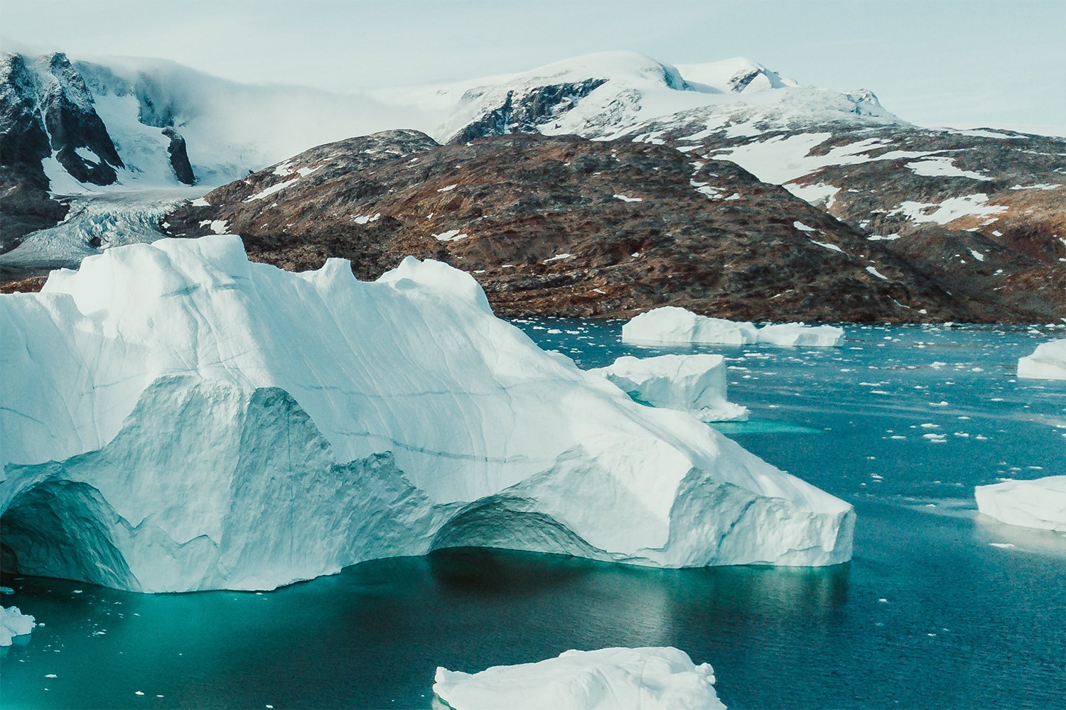 Large icebergs floating in blue water with snow-capped mountains and rocky hills in the background under a cloudy sky.
