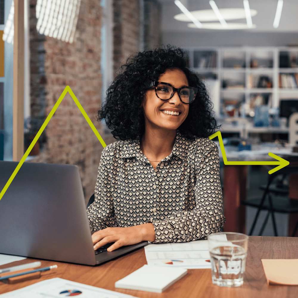A woman with curly hair and glasses smiles while working on a laptop at a desk in a modern office. A yellow arrow graphic is overlaid, suggesting growth or progress. Papers and a glass of water are on the table.