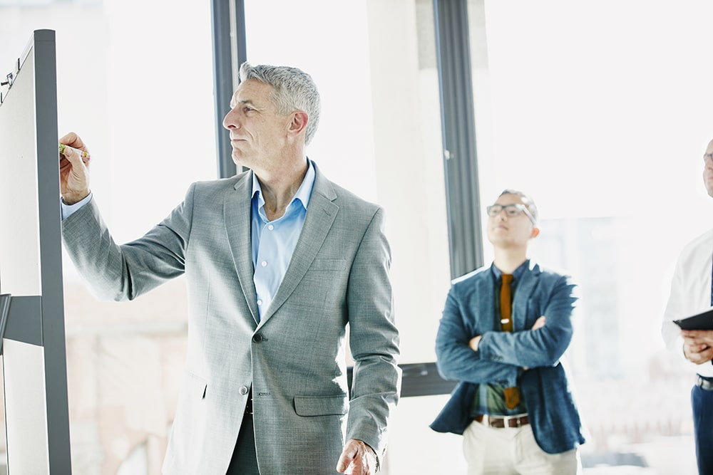 mid-aged man writing on whiteboard; two other men look on 