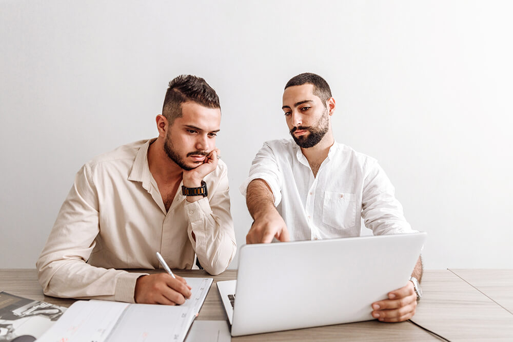 two male presenting coworkers huddling around a laptop at a desk