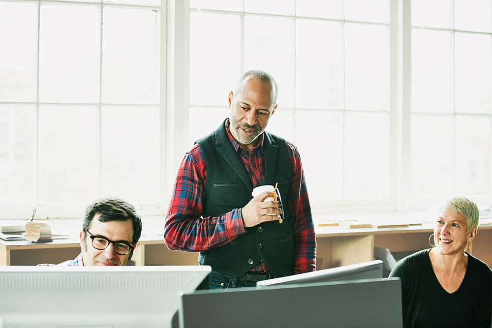 three coworkers, with one man in a plaid shirt and vest overseeing two others 