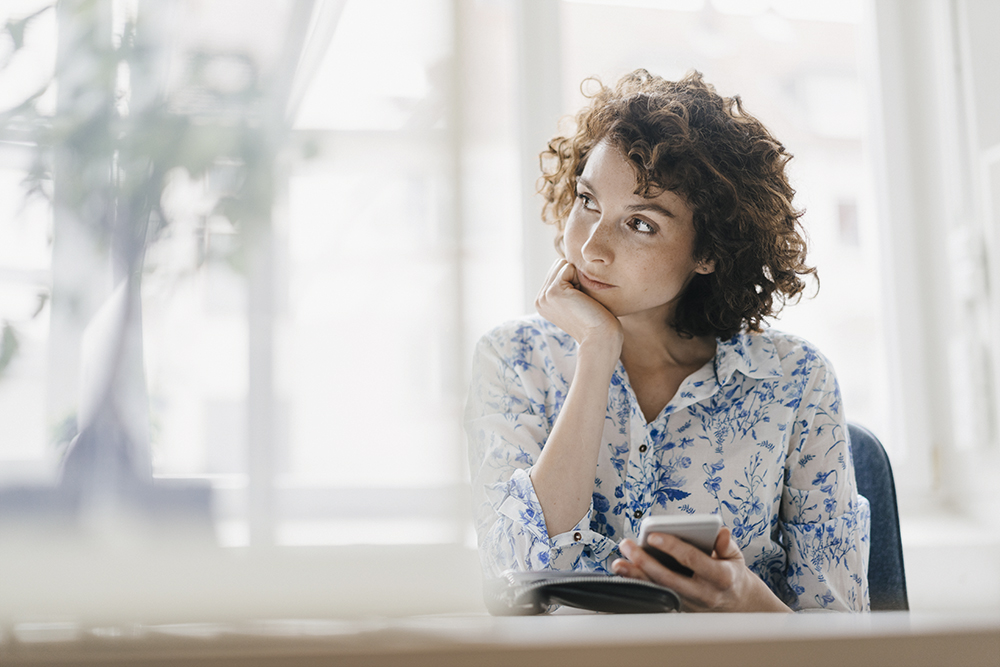 young professional white woman with curly brown hair in flowered shirt, thinking at her desk