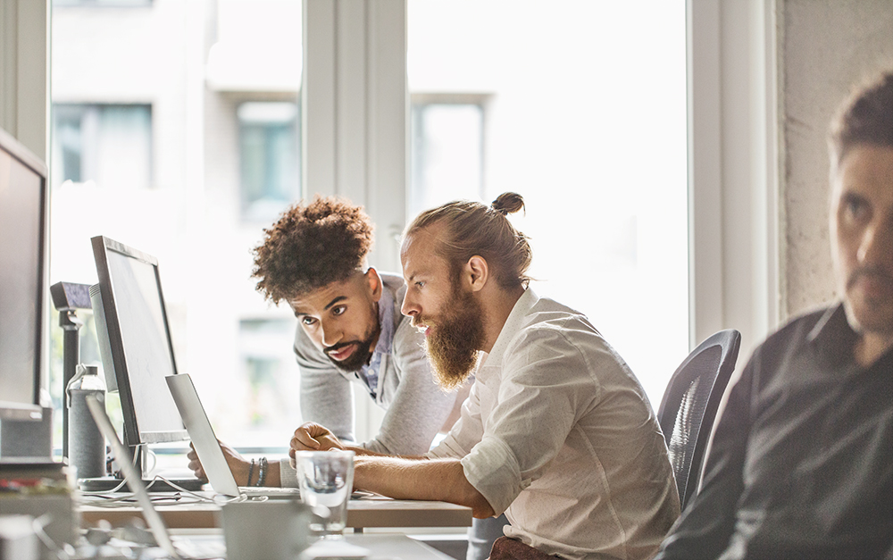 two bearded men working at an office with one other man in the foreground