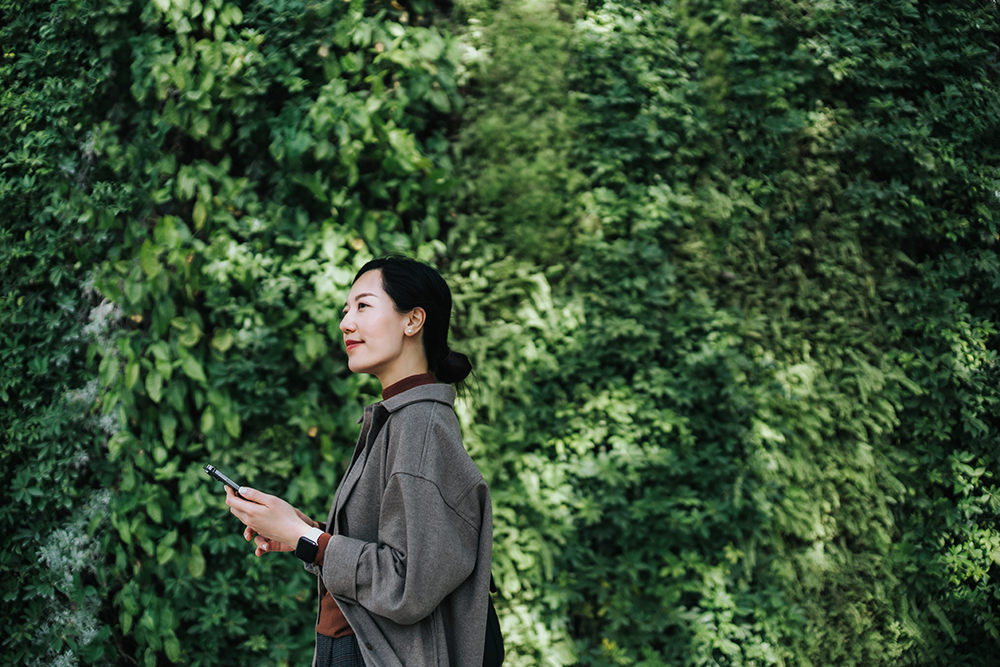 Asian woman walking with phone in front of greenery 