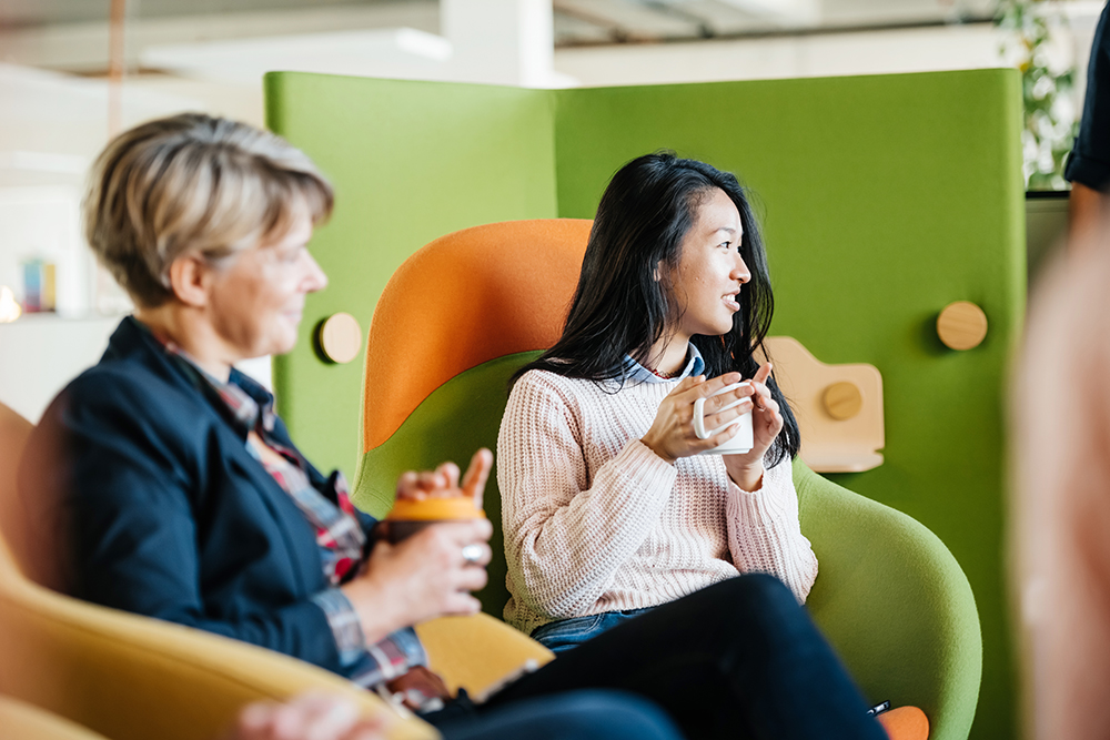 two women holding coffee cups, sitting in green and orange seats in a modern office space talking to other people out of view