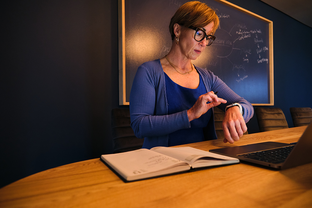 middle aged white woman checking her watch at conference table with laptop and notebook at night