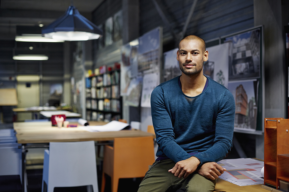 man in blue shirt sitting on the edge of a desk in design agency office with posters and books in the background