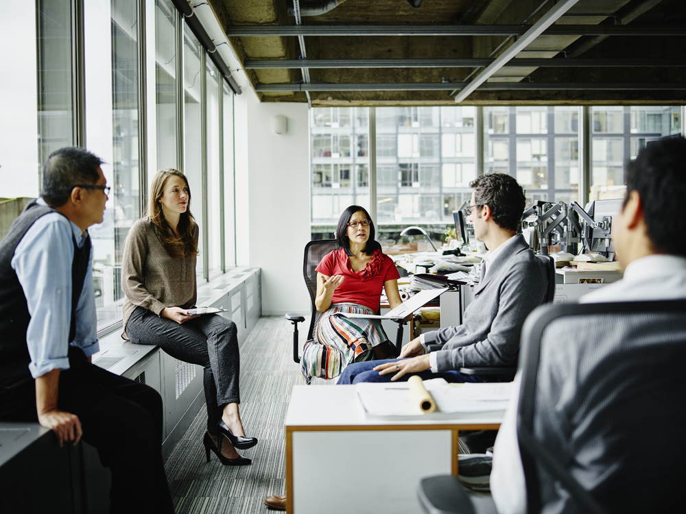A group of five people is engaged in a discussion in a modern office. They are seated around a workspace with large windows in the background, allowing natural light to fill the room. The atmosphere appears collaborative and focused.