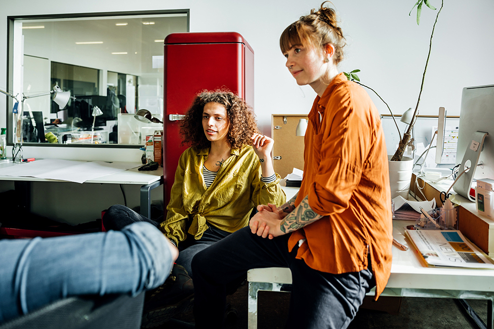 Woman in orange sitting on office desk next to coworker in yellow talking with team