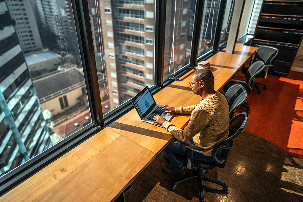 A person sits at a wooden desk working on a laptop in a high-rise office, with large windows offering a view of city buildings. The office is equipped with multiple chairs and desks, and the person is focused on their screen.