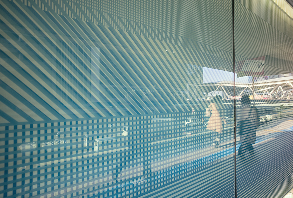 Blue striped glass wall reflecting two people standing at a train station with tracks visible in the background. The stripes create a geometric pattern that partially obscures the view.