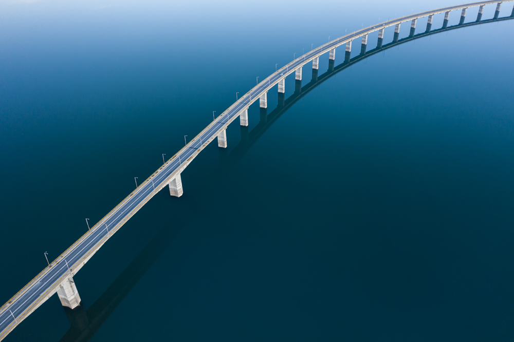 Aerial view of a long, curving bridge spanning across a calm body of water. The bridges reflection creates an elegant arc against the deep blue surface. The scene is serene and expansive, with no vehicles or people visible.