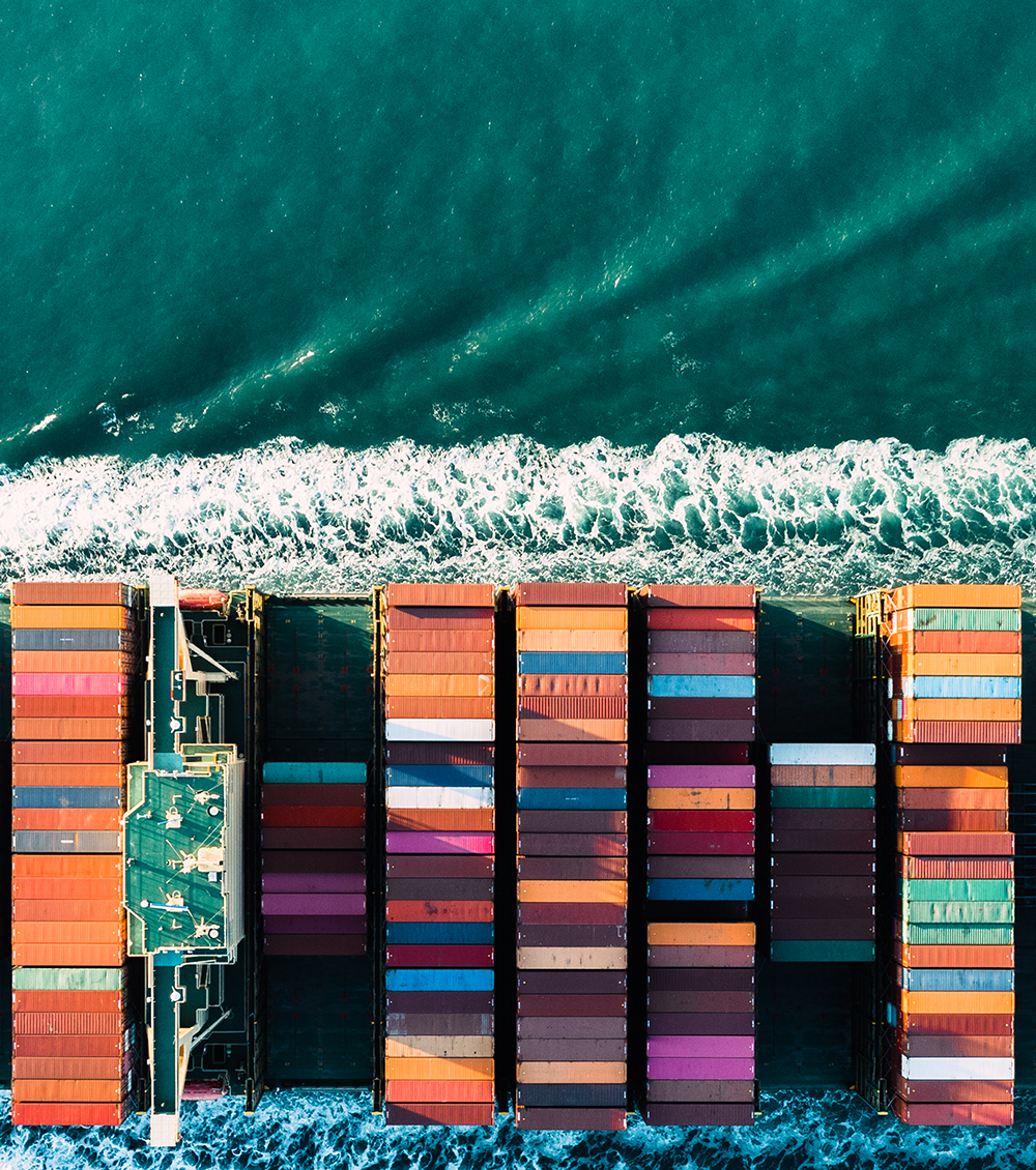Aerial view of a container ship at sea with colorful containers arranged in rows. The turquoise water creates a contrast as the ship cuts through the waves, leaving a trail of white foam behind.