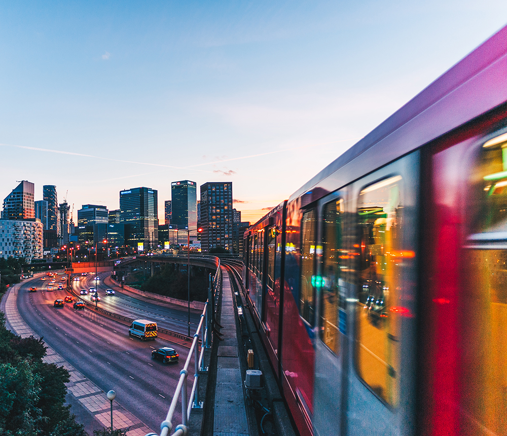 A red train speeds past on an elevated track, overlooking a busy cityscape at sunset. Tall buildings form the skyline, and cars drive on the road below. The sky is a gradient of soft blues and oranges.