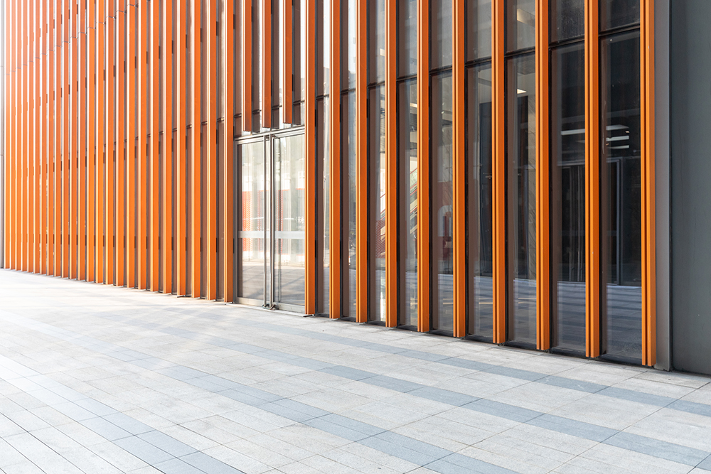 Modern building facade with tall vertical orange slats and reflective glass windows. The exterior has a sleek architectural design, with a spacious, light-colored tiled walkway in the foreground.