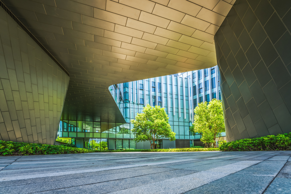 Modern architectural structure with geometric design, featuring a curved metallic canopy. The area below showcases a tiled path and green landscaping with small trees, surrounded by reflective glass buildings. Bright and sunny atmosphere.