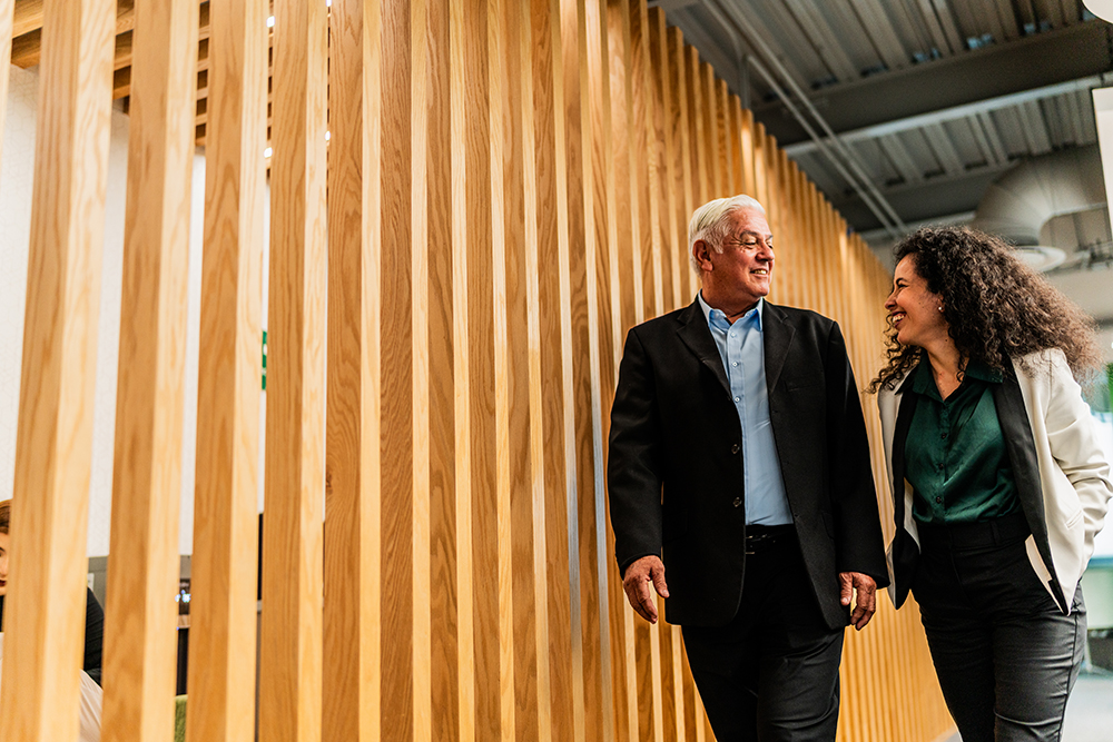 A man and a woman walk side by side in a modern office setting, with wooden slats as a background. The man wears a black suit, and the woman is in a white blazer and green blouse. They are smiling and engaged in conversation.