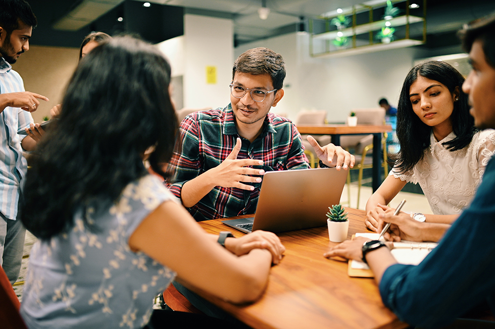 A group of people sitting around a table in a modern office environment. A man with glasses is speaking and gesturing, while others are listening attentively. A laptop and a small plant are on the table. Others are conversing in the background.