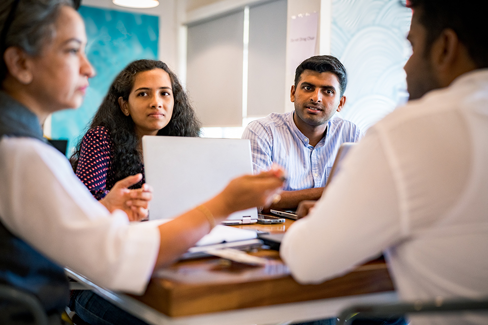 Four people sit together at a table in a modern office setting. Theyre engaged in a discussion, with two laptops visible. A mural is partially visible in the background, creating a colorful ambiance.