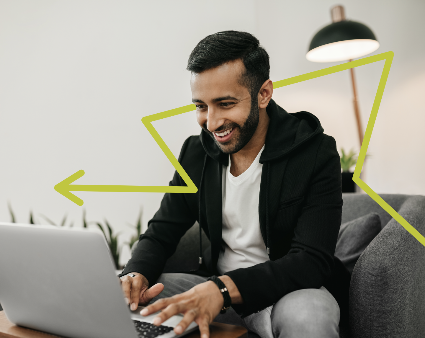 A man with a beard is smiling while using a laptop. Hes sitting on a sofa with plants in the background. A large green zigzag line overlays the image, and a floor lamp is visible behind him.