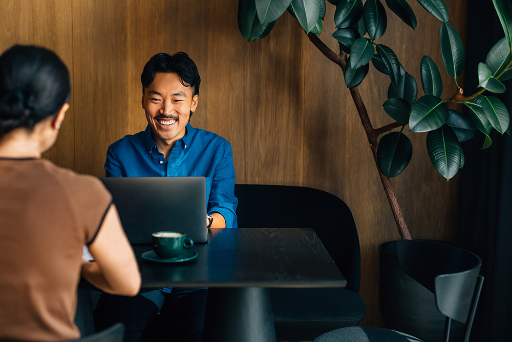 A smiling man in a blue shirt sits at a dark table, using a laptop. Across from him, a woman in a brown shirt is turned away. A cup is on the table. The background features a large green plant against a wooden wall.