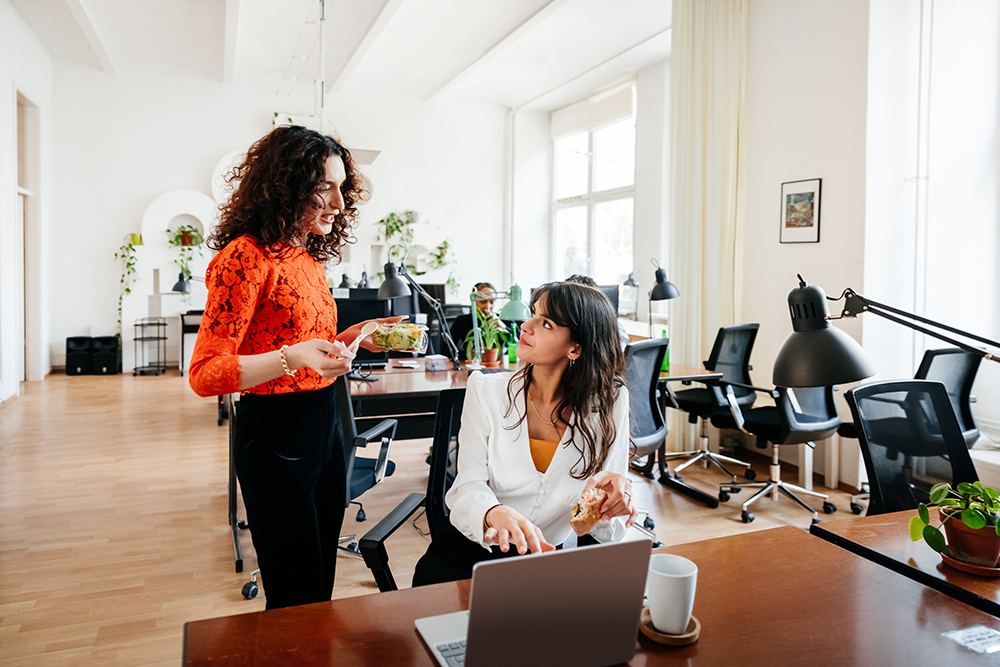 Two women in an office: one standing with a salad, wearing a red top, talking to a seated woman in a white blouse with a sandwich. Open workspace with plants and laptops, wood flooring, and natural light.