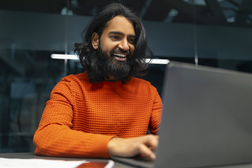 A person with long dark hair and a beard is smiling while working on a laptop. They are wearing an orange sweater and are seated at a desk with papers. The background is blurred with dark tones and lights.