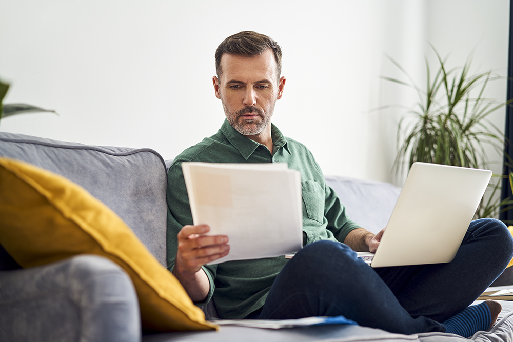 A man sits on a gray sofa holding papers in one hand and a laptop on his lap. He wears a green shirt and jeans. Theres a yellow pillow beside him and a plant in the background, creating a cozy living room ambiance.