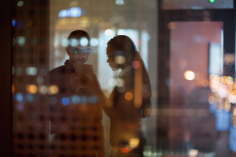 Two people stand indoors at night, looking at a tablet together. Their silhouettes are seen through a glass window, with blurred city lights and reflections creating a colorful, abstract effect.