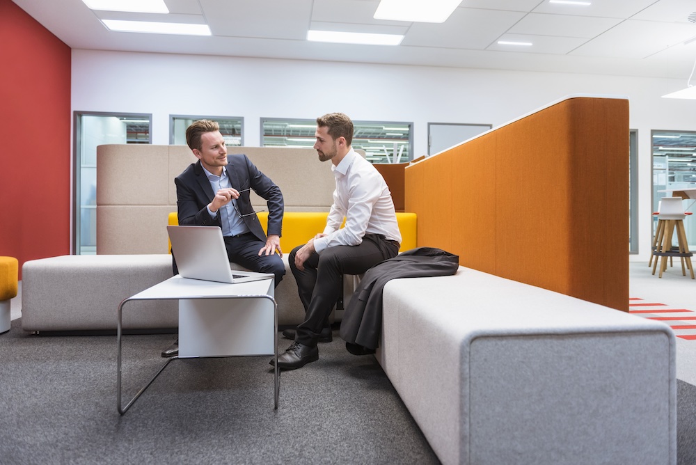 Two men in business attire sit on modern office sofas having a discussion. One gestures while the other listens. A laptop is open on the table between them in a bright, contemporary office space.