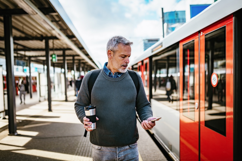 A man with gray hair, wearing a sweater and backpack, stands on a train platform holding a coffee cup and looking at his smartphone, with a red and white train beside him.
