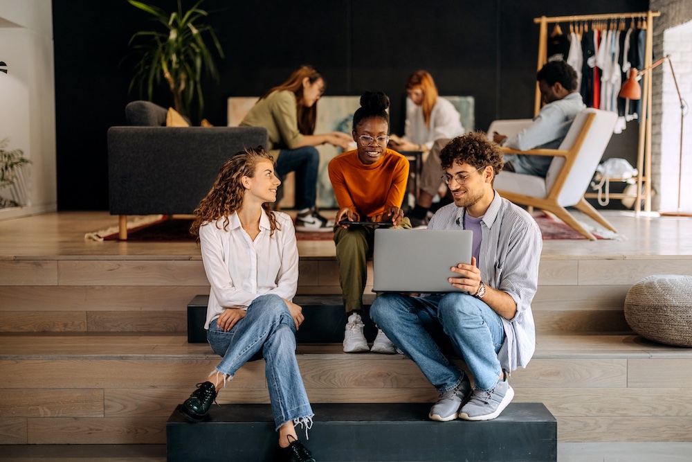A group of young adults sit on steps in a modern, open space. Three people in front smile and look at a laptop, while others in the background chat and work, creating a collaborative atmosphere.