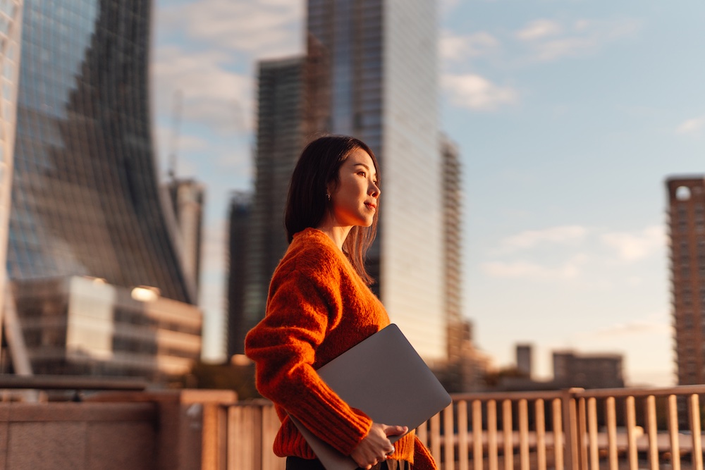 A woman in an orange sweater stands outdoors holding a closed laptop, gazing thoughtfully into the distance with a city skyline and modern tall buildings in the background.