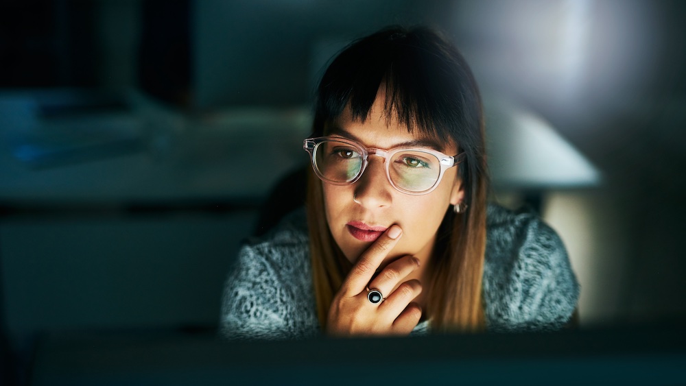 A woman wearing glasses sits in a dimly lit room, looking thoughtfully at a computer screen with her hand resting on her chin.