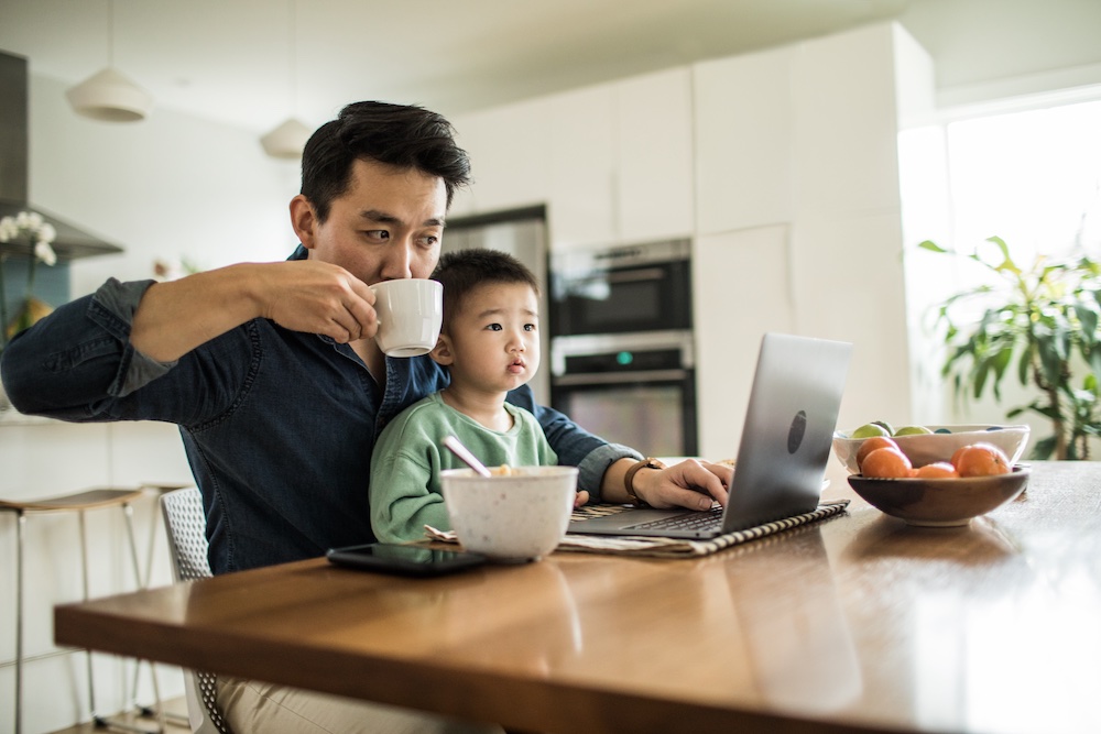 A man drinks from a mug while working on a laptop at a kitchen table with a young child on his lap. A bowl of fruit and a bowl of cereal are on the table in a bright, modern home.