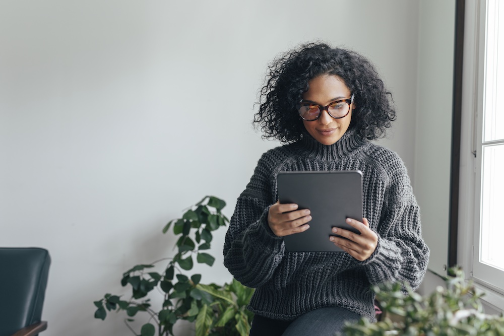 A woman with curly hair and glasses, wearing a gray sweater, sits indoors near a window and plants, holding and looking at a tablet.