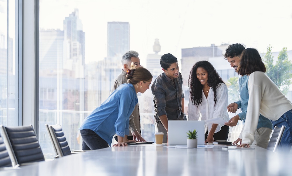 A diverse group of six colleagues stands around a table in a modern office, smiling and discussing something on a laptop. Large windows show a city skyline in the background.