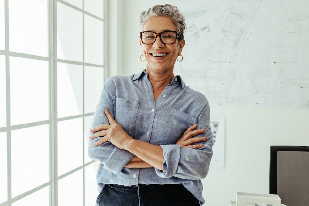 A smiling older woman with short gray hair, wearing glasses, hoop earrings, and a light blue shirt stands with arms crossed in a bright office with architectural plans on the wall behind her.