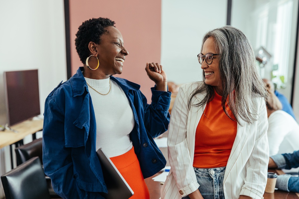 Two women standing and laughing together in an office setting, one holding a laptop. One wears a blue jacket and orange skirt, the other a white blazer and orange top. They appear to be enjoying a friendly conversation.