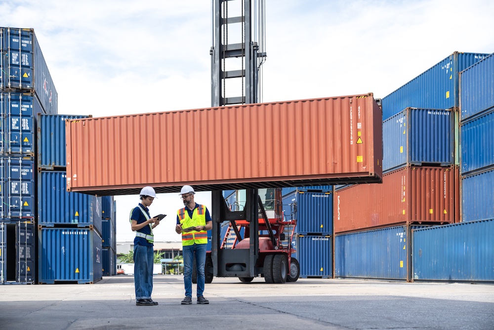 Two workers in safety gear stand in front of stacked shipping containers, discussing on a clipboard, while a forklift hoists an orange container above them in an outdoor shipping yard.